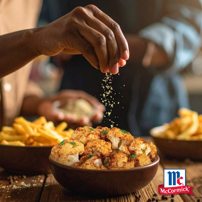Eine Hand streut Gewürze aus dem Mccormick Global Template De Sortiment auf eine Schüssel gerösteten Blumenkohl, mit Pommes frites und einer weiteren Person im Hintergrund.