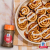 Eine Flasche Srpet Cinnamon Gd X06-Ge steht neben einem Teller mit frisch gebackenen Zimtschnecken mit weißem Zuckerguss auf einer karierten Serviette.