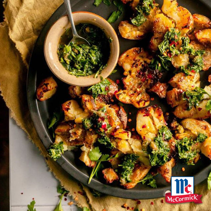 Knusprig zerdrückte Kartoffeln werden mit Srpet Garlic Granul X06-Ge gewürzt und mit einer lebendigen Kräuter-Chimichurri-Sauce auf einer dunklen Platte mit dem McCormick-Logo serviert.