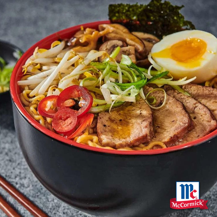 Eine dampfende Schüssel Ramen, belegt mit Schweinefleischscheiben, einem weichgekochten Ei und frischem Gemüse, ist mit Srpet Bl Pepper Gd X06-Ge gewürzt und zeigt das McCormick-Logo.
