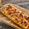 Eine lange rechteckige Fladenbrot-Pizza, belegt mit gegrillten Steakstreifen, geschmolzenem Käse, gewürfelten Tomaten und roten Zwiebeln, ist mit Srpet Pizza Seas X06-Ge gewürzt und wird auf einem rustikalen Holzbrett serviert.