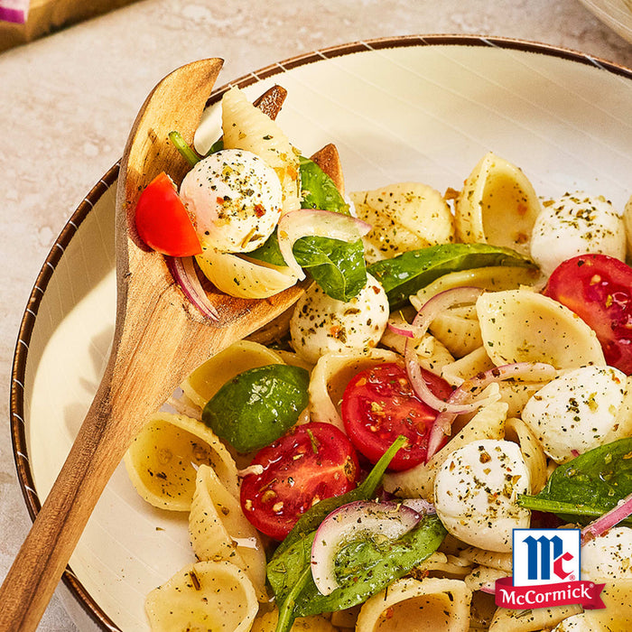 Ein Holzlöffel hebt eine Portion Nudelsalat mit Orecchiette, frischem Basilikum, roten Zwiebeln, Kirschtomaten und Mozzarella-Perlen, die mit Srpet Tomato Mozza X06-Ge und dem McCormick-Logo in der Ecke gewürzt sind.