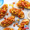 Eine Marmoroberfläche beherbergt mehrere Stücke geröstetes Brot, die mit einer farbenfrohen Mischung aus roten und gelben Tomaten belegt sind, die mit Srpet Tomato Mozza X06-Ge und dem McCormick-Logo in der Ecke gewürzt sind.