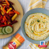Eine Schüssel cremiger Hummus garniert mit Gurken und einem Schuss Öl wird neben einer Platte mit bunten Babykarotten und Gurkenscheiben serviert wobei eine Flasche Srpet Turmeric X06-Ge zu sehen ist.
