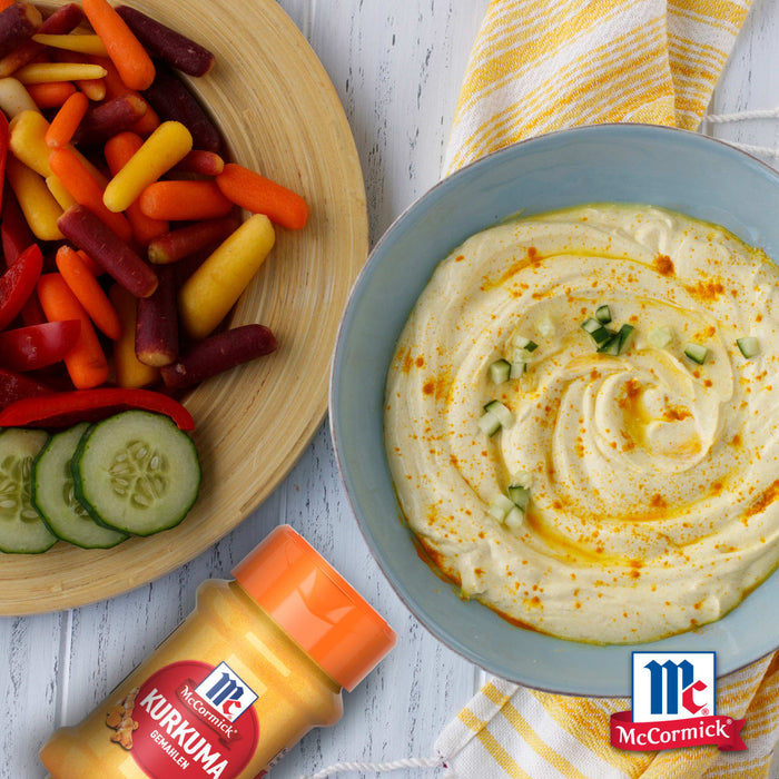 Eine Schüssel cremiger Hummus garniert mit Gurken und einem Schuss Öl wird neben einer Platte mit bunten Babykarotten und Gurkenscheiben serviert wobei eine Flasche Srpet Turmeric X06-Ge zu sehen ist.
