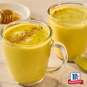 Zwei Glastassen gefüllt mit einem warmen Golden Milk Latte aus Srpet Turmeric X06-Ge sind mit einer Prise Gewürz garniert und neben einem Honiglöffel und dem McCormick Logo abgebildet.